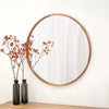 Round mirror with copper frame on a white wall above a wooden surface with two dark glass vases and red dried flowers.