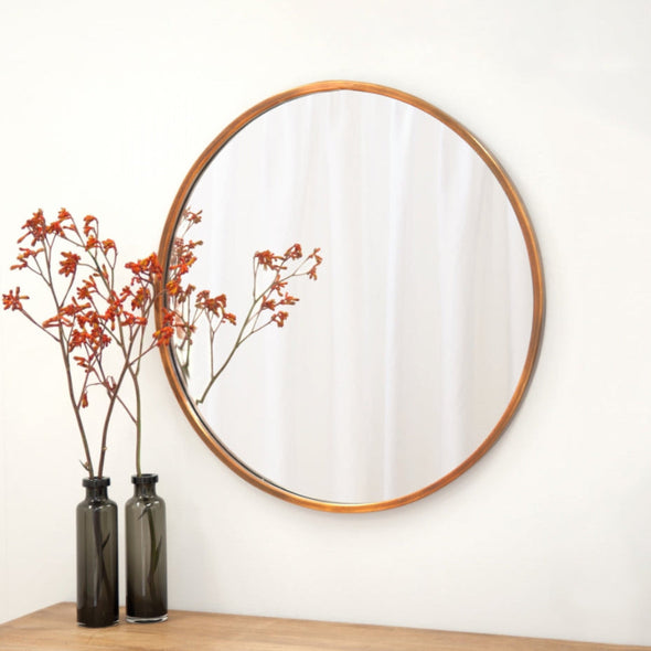 Round mirror with copper frame on a white wall above a wooden surface with two dark glass vases and red dried flowers.