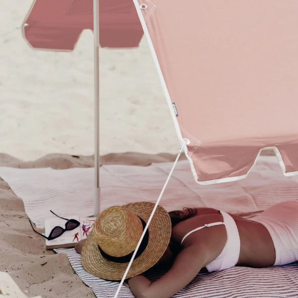Person relaxing under a pink beach umbrella on a sandy beach.