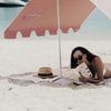 Woman reading a book under a pink beach umbrella on a sandy beach.