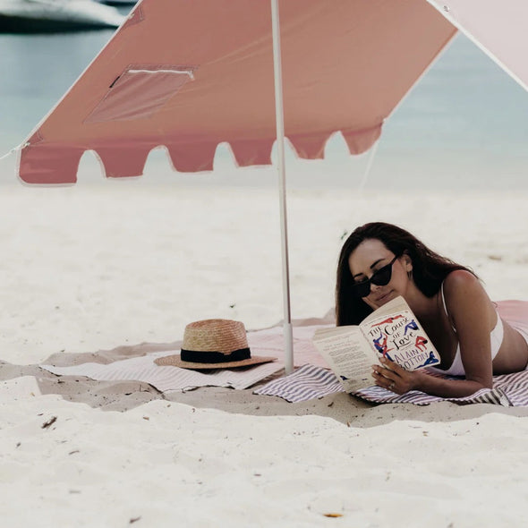 Woman reading a book under a pink beach umbrella on a sandy beach.
