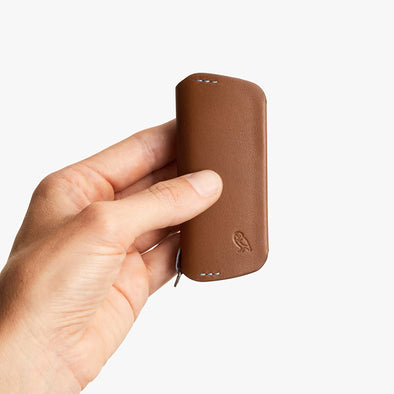 Hand holding a brown leather phone case with a brand logo on a white background