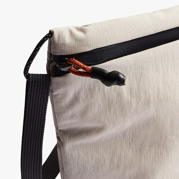 Close-up of a beige bag with a black strap and orange zipper pull on a white background