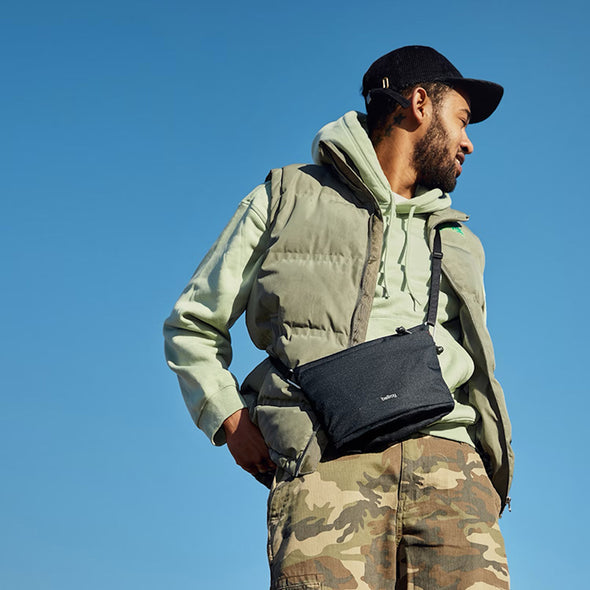 Man wearing a green jacket and camouflage pants with a black bag against a clear blue sky.