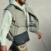 Person wearing a light green puffer vest and holding a black crossbody bag against a neutral background