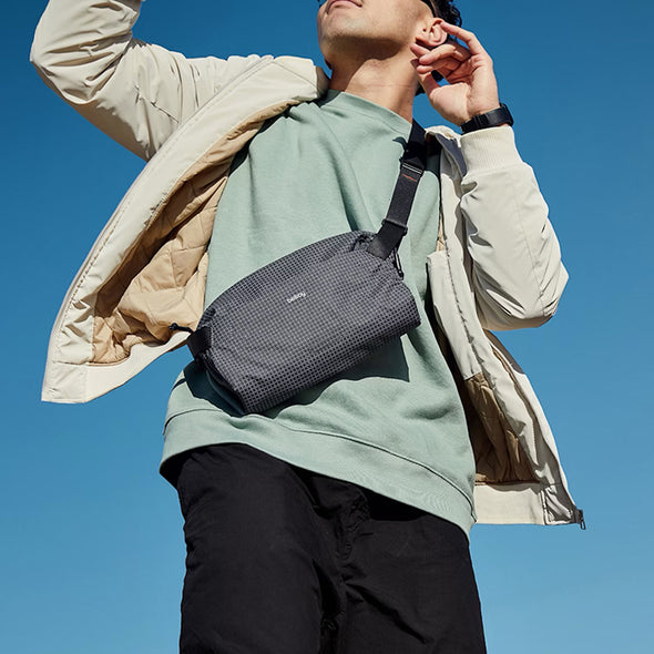 Person wearing a green shirt and black pants with a gray crossbody bag against a clear blue sky.