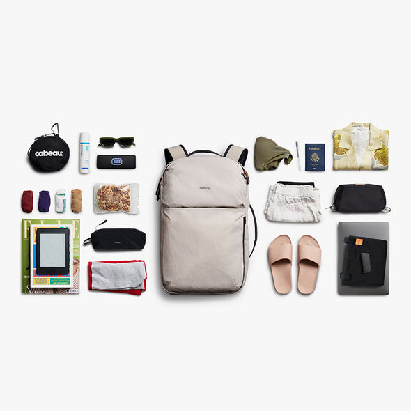 Collection of travel items arranged around a beige backpack on a white background
