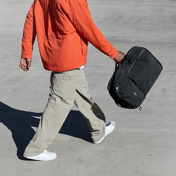 Person in orange jacket and beige pants carrying a black bag on a gray pavement.