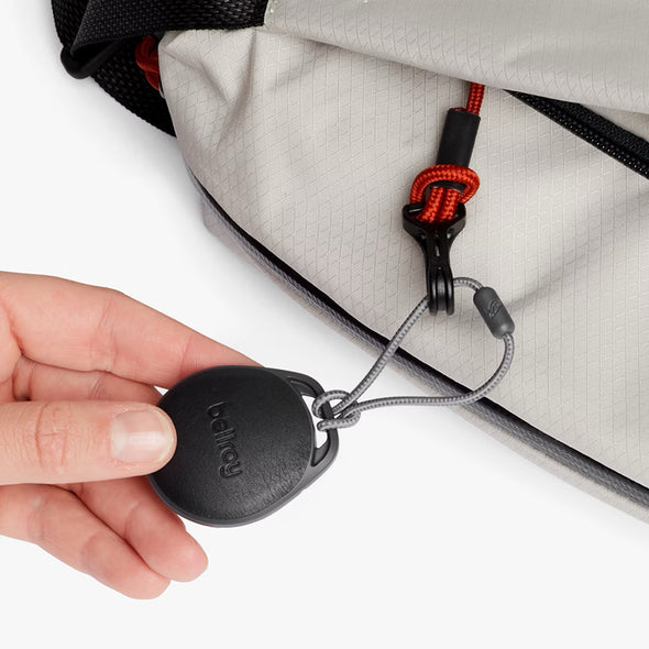 Hand holding a black device with a red and black lanyard attached to a gray bag.