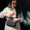 Woman in a gray coat and sunglasses holding a black bag and brown wallet against a dark background