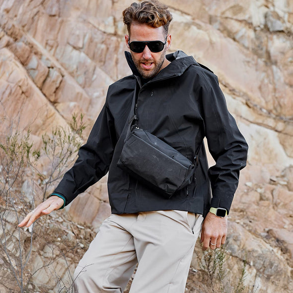 Man wearing a black jacket and sunglasses with a black bag, standing against a rocky background.