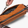 Brown crossbody bag with a hand placing keys into it on a light gray background