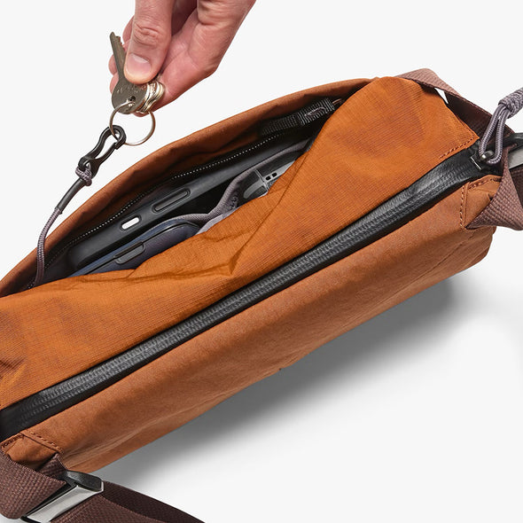 Brown crossbody bag with a hand placing keys into it on a light gray background