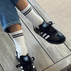 Person wearing black Adidas sneakers with white socks on a wooden floor.