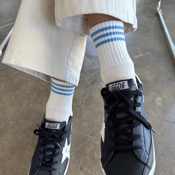 Person wearing black sneakers with 'GGDB' branding and white socks with blue stripes on a concrete floor.