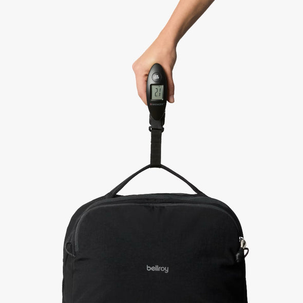Black backpack with a digital luggage scale attached, held by a hand against a white background.