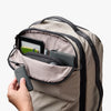 Beige backpack with a hand reaching in to open a compartment, showing a tablet and phone.