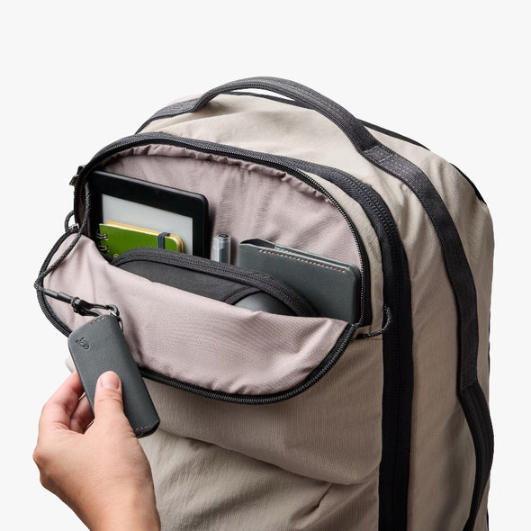Beige backpack with a hand reaching in to open a compartment, showing a tablet and phone.