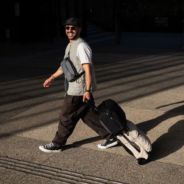 Man walking with a suitcase on a street, wearing sunglasses and a vest.