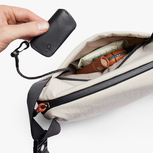 Hand holding a black key case above an open beige bag with travel items.