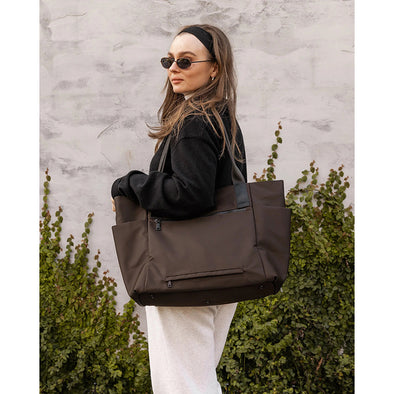 Woman holding a brown tote bag against a textured wall and green plants background