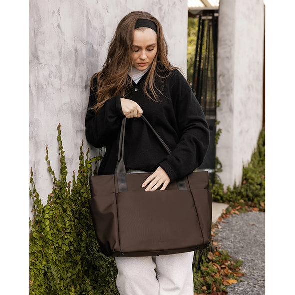 Woman holding a brown tote bag against a light-colored wall with greenery.