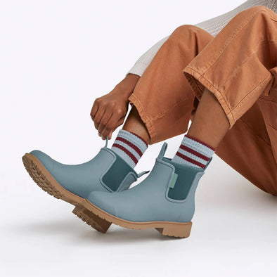 Light blue rain boots with brown soles worn by a person in brown pants and striped socks.