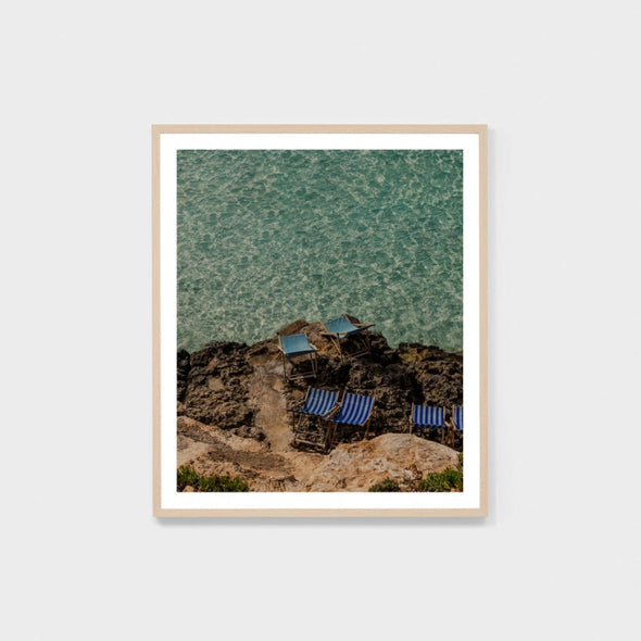 Framed artwork of lounge chairs on a rocky beach with turquoise water.