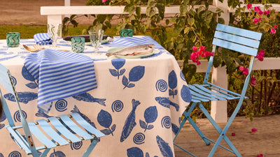 Outdoor setting with a table set for a meal on a patterned tablecloth, surrounded by greenery and flowers.