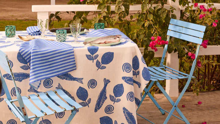 Outdoor setting with a table set for a meal on a patterned tablecloth, surrounded by greenery and flowers.