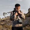 Man in outdoor setting wearing a beige jacket and black beanie, holding a black bag.