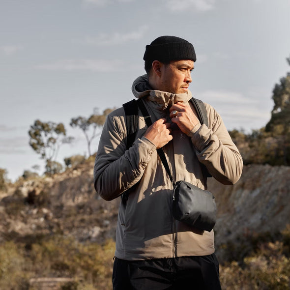 Man in outdoor setting wearing a beige jacket and black beanie, holding a black bag.