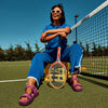 Person sitting on a tennis court with a racket, wearing sunglasses and a blue outfit.