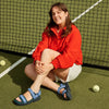 Woman in red jacket and blue sandals sitting on a tennis court with tennis balls around.