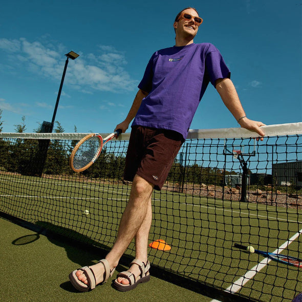 Man in purple shirt and sunglasses holding a tennis racket on a tennis court.