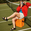 Person sitting on an orange cooler taking a photo with a vintage camera on a tennis court.